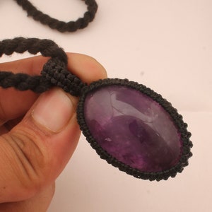 May include: A purple amethyst gemstone pendant on a black cord necklace. The pendant is oval shaped and has a smooth, polished surface.