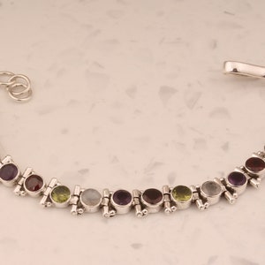 Könnte beinhalten: Ein silbernes Armband mit einem Kettenverschluss und einer Reihe von abwechselnden Edelsteinen in Violett-, Grün- und Weißtönen.