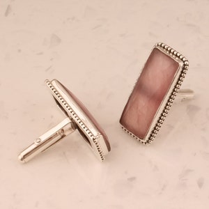May include: A pair of silver cufflinks with a pink rectangular stone set in each. The stones are surrounded by a beaded border.