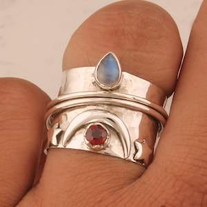 May include: A silver spinner ring with a hammered texture. The ring features a teardrop-shaped, light blue gemstone and a round, red gemstone. The design includes a crescent moon and star cutouts.