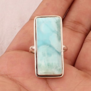 Könnte beinhalten: Ein rechteckiger Silberring mit einem großen, hellblauen Edelstein mit weißen Marmorierungen. Der Edelstein ist in einem einfachen, polierten Silberband gefasst. Der Ring wird auf einer Hand präsentiert.