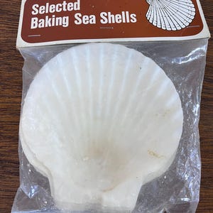 May include: A packet of selected baking sea shells. The shells are white and in a clear plastic bag. The package has a brown label with the words "Selected Baking Sea Shells" in white text.