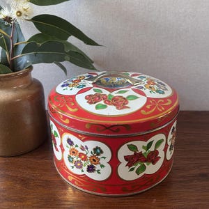 May include: A round red metal tin featuring a white floral pattern with red roses and multi-coloured flowers. The lid has a silver-toned metal handle attached to the centre. The sides of the tin are decorated with gold-coloured scrollwork accents.