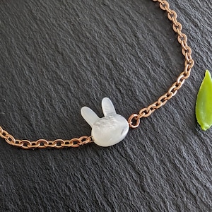 Può includere: Bracciale con catena in oro rosa con un ciondolo a forma di coniglietto bianco. Il ciondolo a forma di coniglietto ha una superficie strutturata. Una piccola foglia verde è accanto al bracciale su una superficie scura. Il bracciale è un gioiello delicato.