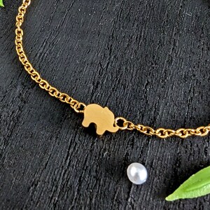 Può includere: Un braccialetto a catena dorata con un piccolo ciondolo a forma di elefante. Il braccialetto è su una superficie nera con una perla bianca e foglie verdi.