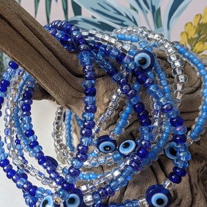Peut inclure: Un ensemble de bracelets en perles bleues et transparentes avec des breloques en forme d'œil maléfique. Les bracelets sont empilés les uns sur les autres et sont faits de petites perles rondes.