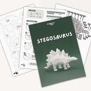 May include: A papercraft kit for building a Stegosaurus dinosaur. The kit includes instructions, a diagram, and a green background with the title "kumera. kit STEGOSAURUS".
