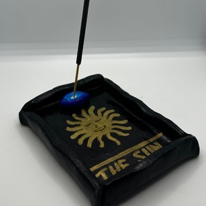 May include: Black rectangular incense holder with a gold sun design and the words "THE SUN". A lit incense stick stands upright in the holder. The holder has a blue and gold accent.