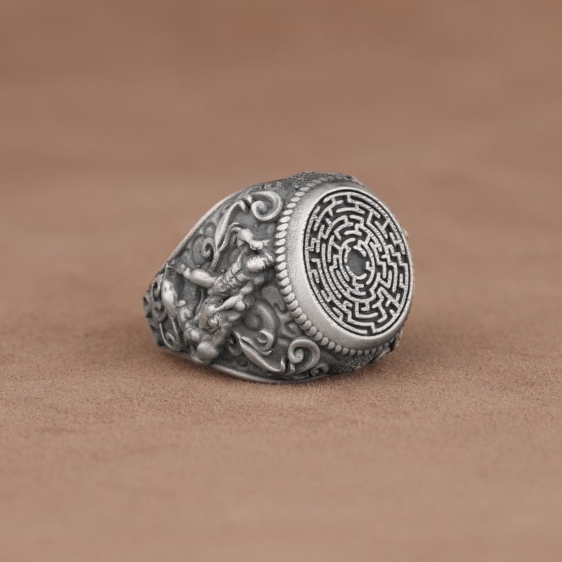 May include: A silver ring with a detailed design featuring a mythical creature and a circular maze pattern on the top.