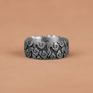 Può includere: Un anello d'argento con un design dettagliato di nuvole vorticose. L'anello è largo e ha una superficie testurizzata.