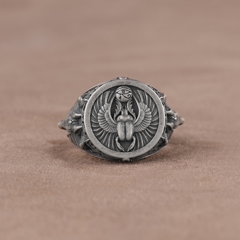 May include: A silver ring with a detailed design of an Egyptian scarab beetle with wings and the Eye of Horus. The ring has a textured band.