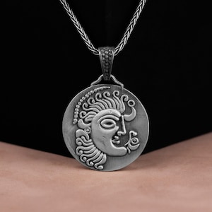 May include: Silver necklace with a round pendant featuring a detailed embossed design of a face with curly hair.