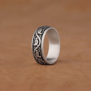 Puede incluir: Un anillo de plata con un patr&oacute;n decorativo de p&aacute;jaros y vides estilizados alrededor de la banda.