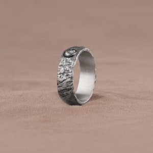 May include: A silver ring with a textured surface and a small, round, black stone inset. The ring is lying on a brown surface.