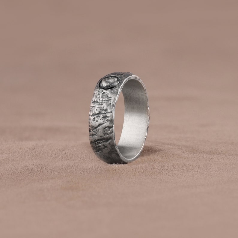 May include: A silver ring with a textured surface and a small, round, black stone inset. The ring is lying on a brown surface.