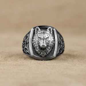 May include: A silver ring with a wolf head design. The wolf's head is detailed and realistic, with a textured fur pattern. The ring band is decorated with intricate floral designs.