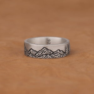 Puede incluir: Un anillo de plata con un diseño de cordillera y un pequeño árbol en el lateral. El anillo está marcado con "92" en el interior.