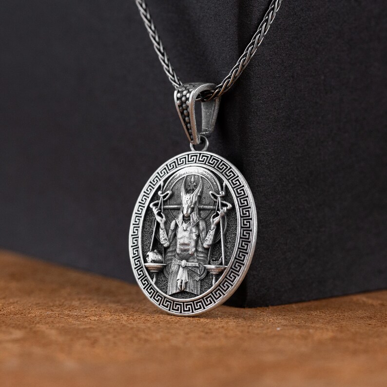 May include: Silver pendant necklace featuring a detailed depiction of a horned figure holding scales, surrounded by a Greek key border.
