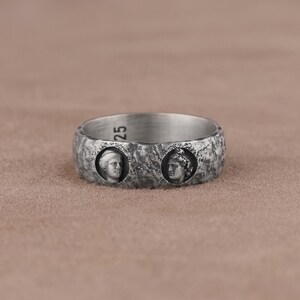 May include: A silver ring with two profile portraits of people with a textured surface.