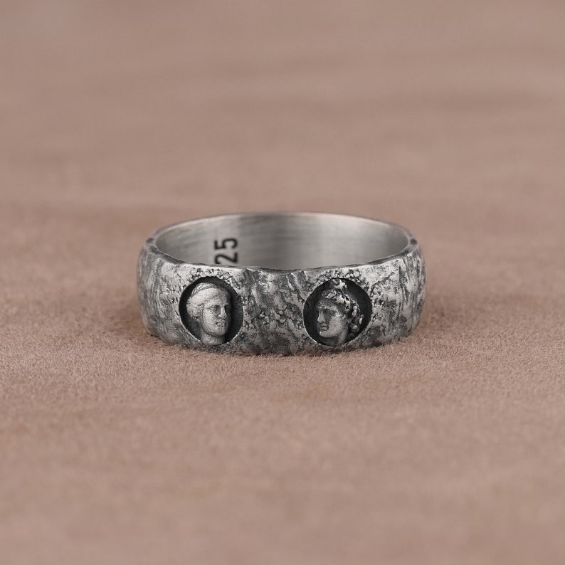 May include: A silver ring with two profile portraits of people with a textured surface.