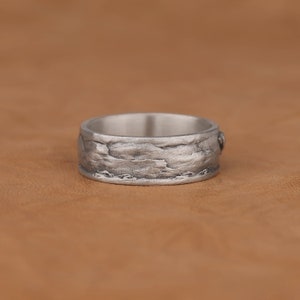 May include: A silver ring with a textured, rocky design. The ring is wide and has a smooth inner band.