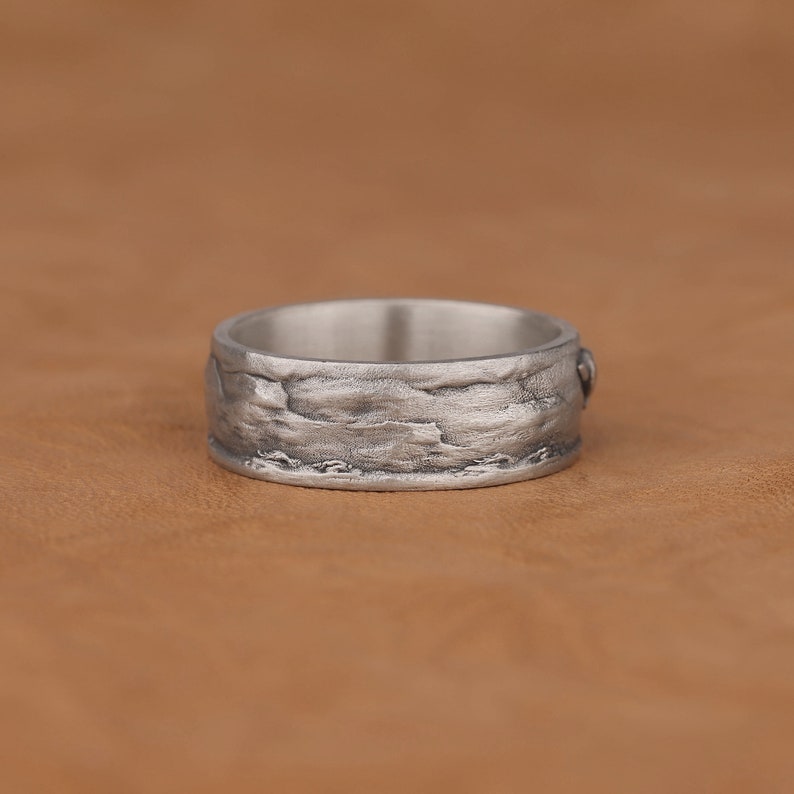May include: A silver ring with a textured, rocky design. The ring is wide and has a smooth inner band.