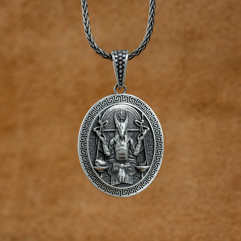 May include: A silver pendant necklace featuring a detailed design of a figure holding scales. The pendant is oval shaped and has a Greek key border.