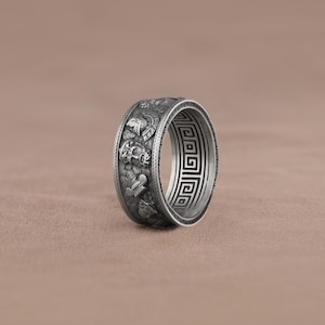 K&ouml;nnte beinhalten: Ein silberner Ring mit einem detaillierten Design von griechischen Kriegern und einem griechischen Schl&uuml;sselmuster um das Band.