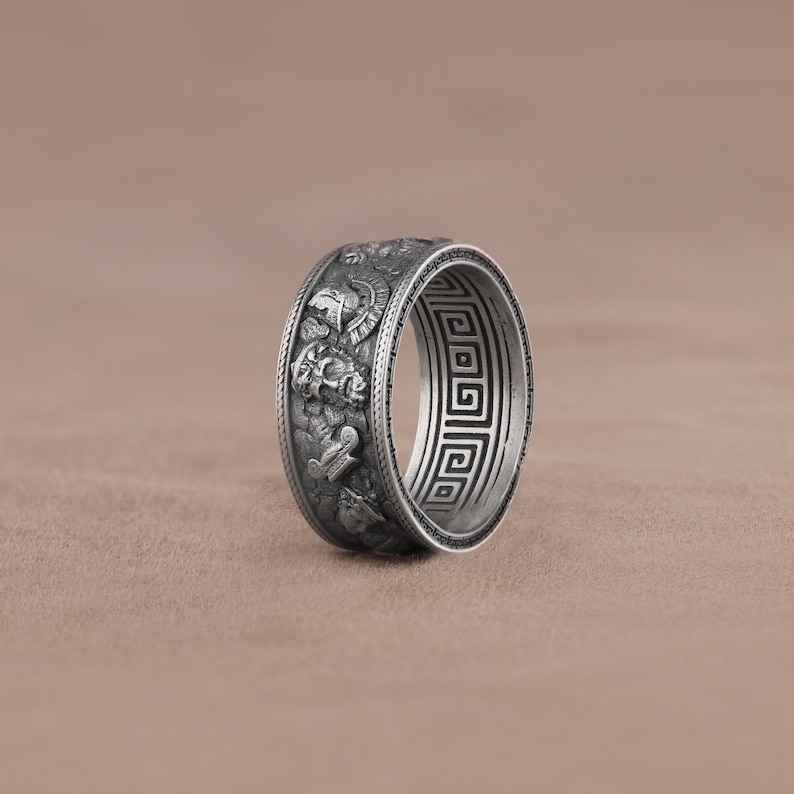 K&ouml;nnte beinhalten: Ein silberner Ring mit einem detaillierten Design von griechischen Kriegern und einem griechischen Schl&uuml;sselmuster um das Band.