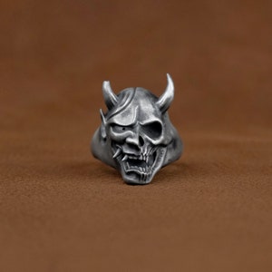 May include: A silver ring with a detailed design of a demon mask. The mask has sharp horns, a fierce expression, and a skull-like face.