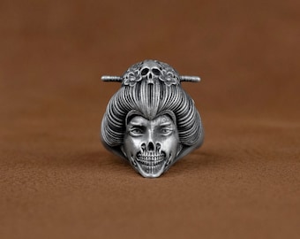May include: A silver ring featuring a detailed skull face design. The skull is wearing a traditional Japanese geisha hairstyle with a flower ornament.