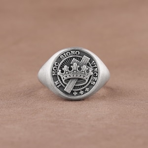 Könnte beinhalten: Ein silberner Siegelring mit einem erhabenen Design, das eine Krone, ein Kreuz und den lateinischen Spruch "In Hoc Signo Vinces" zeigt.