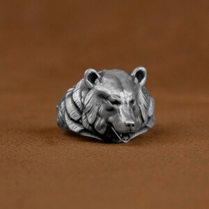 May include: A silver ring with a detailed bear head design. The bear's mouth is slightly open, showing its teeth. The ring is made of metal and has a textured surface.