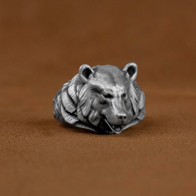 May include: A silver ring with a detailed bear head design. The bear's mouth is slightly open, showing its teeth. The ring is made of metal and has a textured surface.