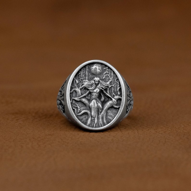 May include: A silver ring featuring an intricate design of a woman standing in a forest with a full moon above her. The ring is oval shaped and has a textured finish.
