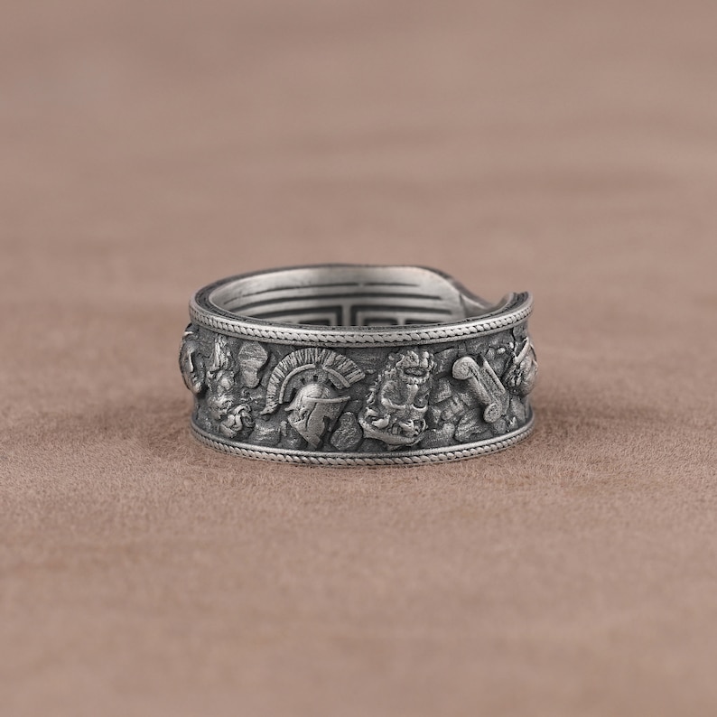 K&ouml;nnte beinhalten: Ein silberner Ring mit einem komplizierten Design, das verschiedene Symbole zeigt, darunter einen Helm, einen L&ouml;wen und eine Schriftrolle. Der Ring ist breit und hat eine strukturierte Oberfl&auml;che.