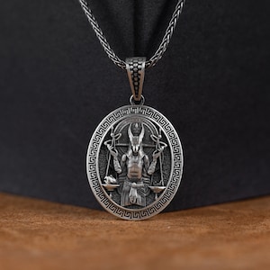 May include: Silver pendant necklace featuring a detailed depiction of a horned figure holding scales, surrounded by a Greek key border.