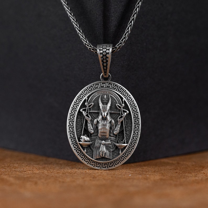 May include: Silver pendant necklace featuring a detailed depiction of a horned figure holding scales, surrounded by a Greek key border.