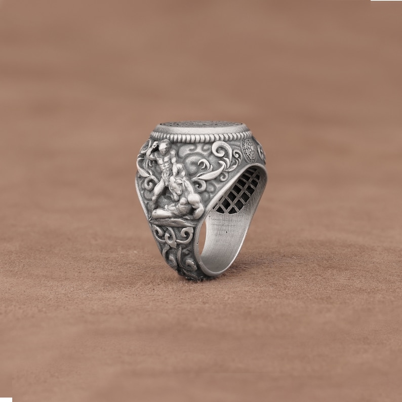 May include: Silver ring with an intricate design featuring two figures wrestling. The ring has a wide band and a detailed, textured surface.
