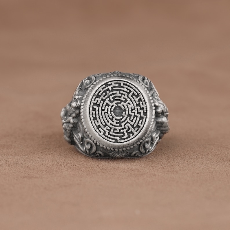 May include: A silver ring with a detailed design featuring a circular maze pattern in the center. The ring has intricate details on the sides.