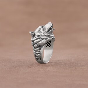 May include: A silver ring with a detailed wolf head design. The wolf's mouth is open and its fur is sculpted in detail. The ring band is textured and has a geometric pattern.