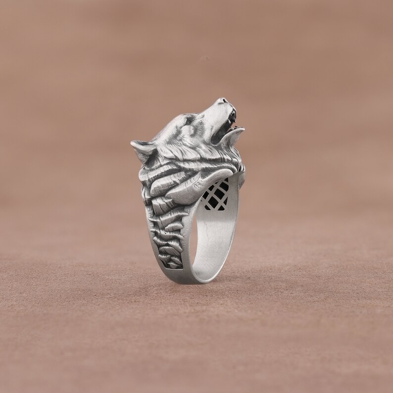 May include: A silver ring with a detailed wolf head design. The wolf's mouth is open and its fur is sculpted in detail. The ring band is textured and has a geometric pattern.