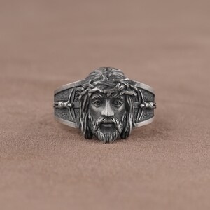 May include: A silver ring with a detailed depiction of Jesus' face, wearing a crown of thorns. The ring features a cross design on the band.