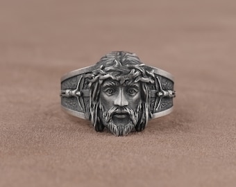 May include: A silver ring with a detailed depiction of Jesus' face, wearing a crown of thorns. The ring features a cross design on the band.
