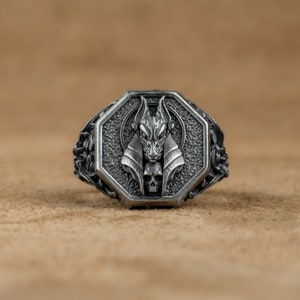 Puede incluir: Un anillo de plata con un diseño octogonal que presenta una representación detallada de una cabeza de Anubis con una calavera debajo.