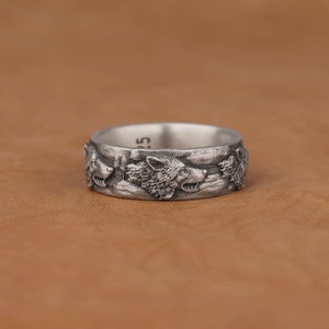 May include: A silver ring with a design of three wolf heads facing forward. The ring is on a brown leather surface.