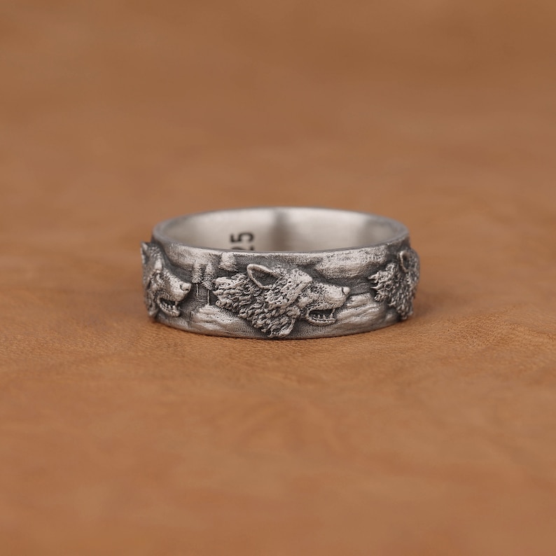 May include: A silver ring with a design of three wolf heads facing forward. The ring is on a brown leather surface.