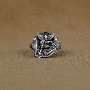 Peut inclure: Une bague en argent avec un motif détaillé d'une moto et d'un aigle. La bague est placée sur un fond brun.