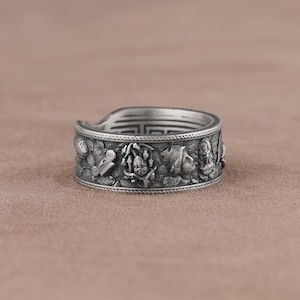 K&ouml;nnte beinhalten: Ein silberner Ring mit einem detaillierten Design, das eine Reihe von erhabenen Figuren und Symbolen zeigt. Der Ring ist breit und hat eine strukturierte Oberfl&auml;che.