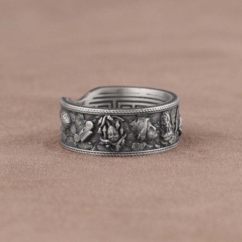 K&ouml;nnte beinhalten: Ein silberner Ring mit einem detaillierten Design, das eine Reihe von erhabenen Figuren und Symbolen zeigt. Der Ring ist breit und hat eine strukturierte Oberfl&auml;che.